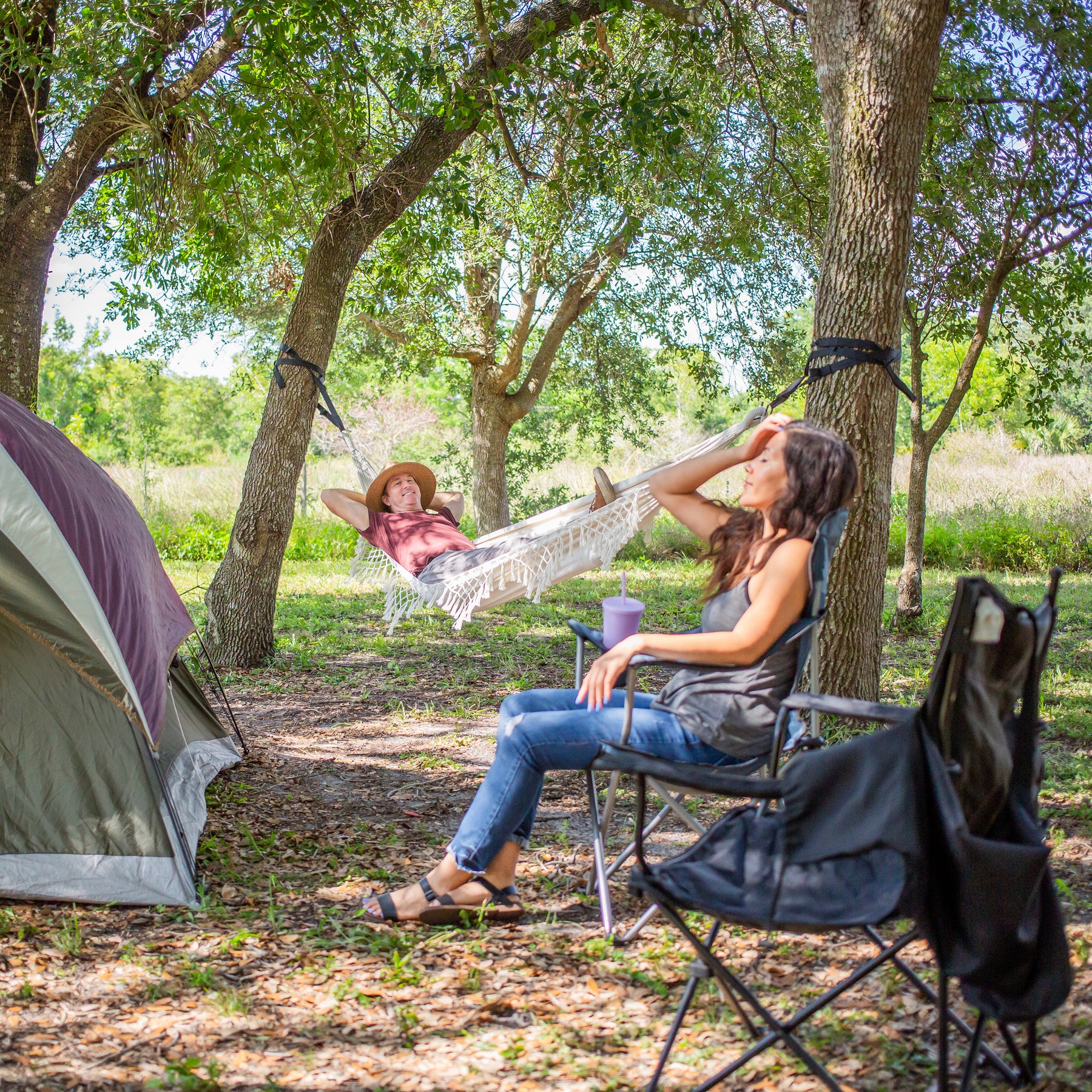 Man laying in the Bliss Hammocks 40-inch Wide Hammock in a Bag with Fringe hung between 2 trees. A woman is sitting in a folding chair in front of him facing a camping tent.