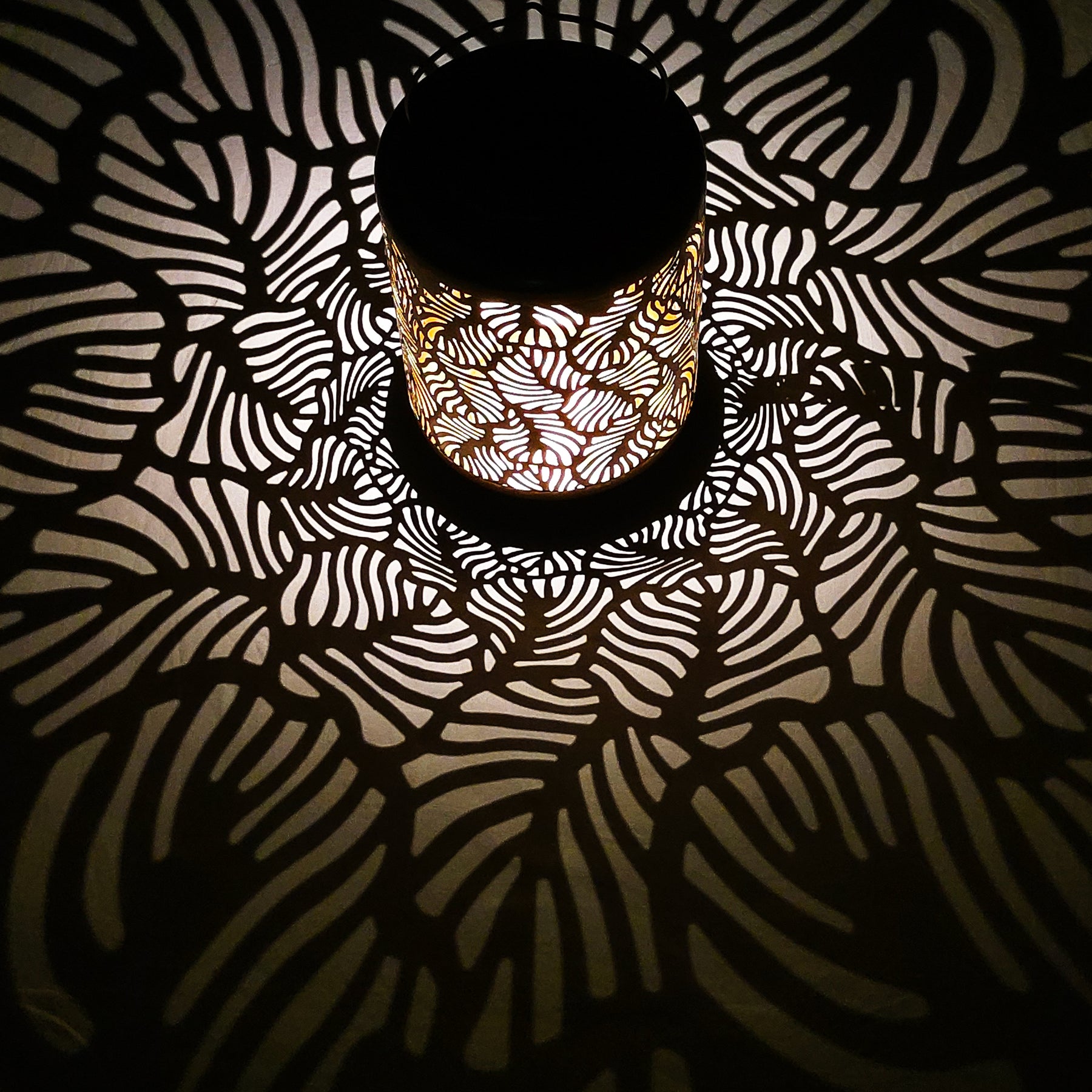 Bliss Outdoors 12-inch Tall Hanging and Tabletop Decorative Solar LED Lantern with Unique Tropical Leaf Design turned on and creating a cool pattern of light on the surface around it.