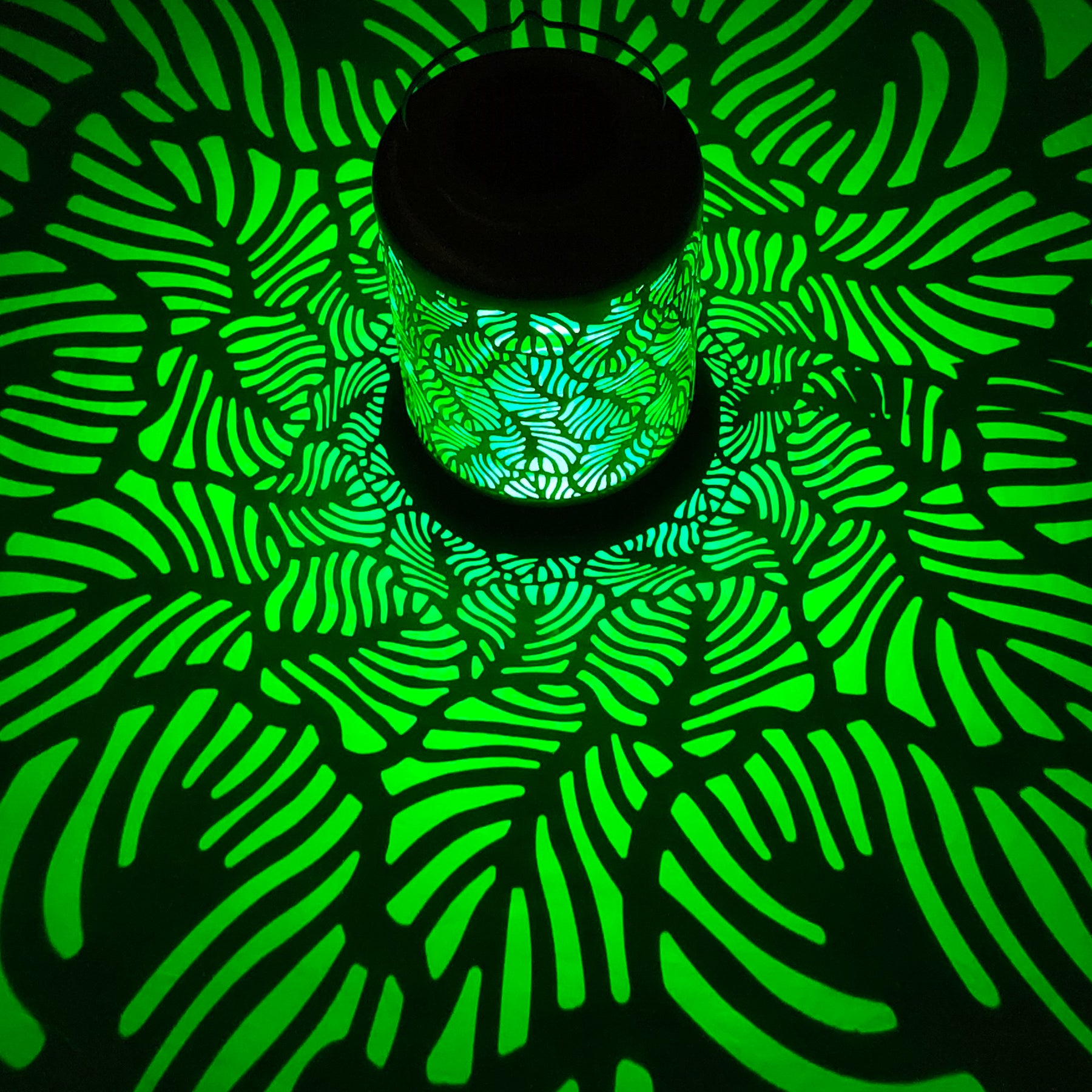 Bliss Outdoors 12-inch Tall Hanging and Tabletop Decorative Solar LED Lantern with Unique Tropical Leaf Design turned on and creating a cool pattern of green light on the surface around it.