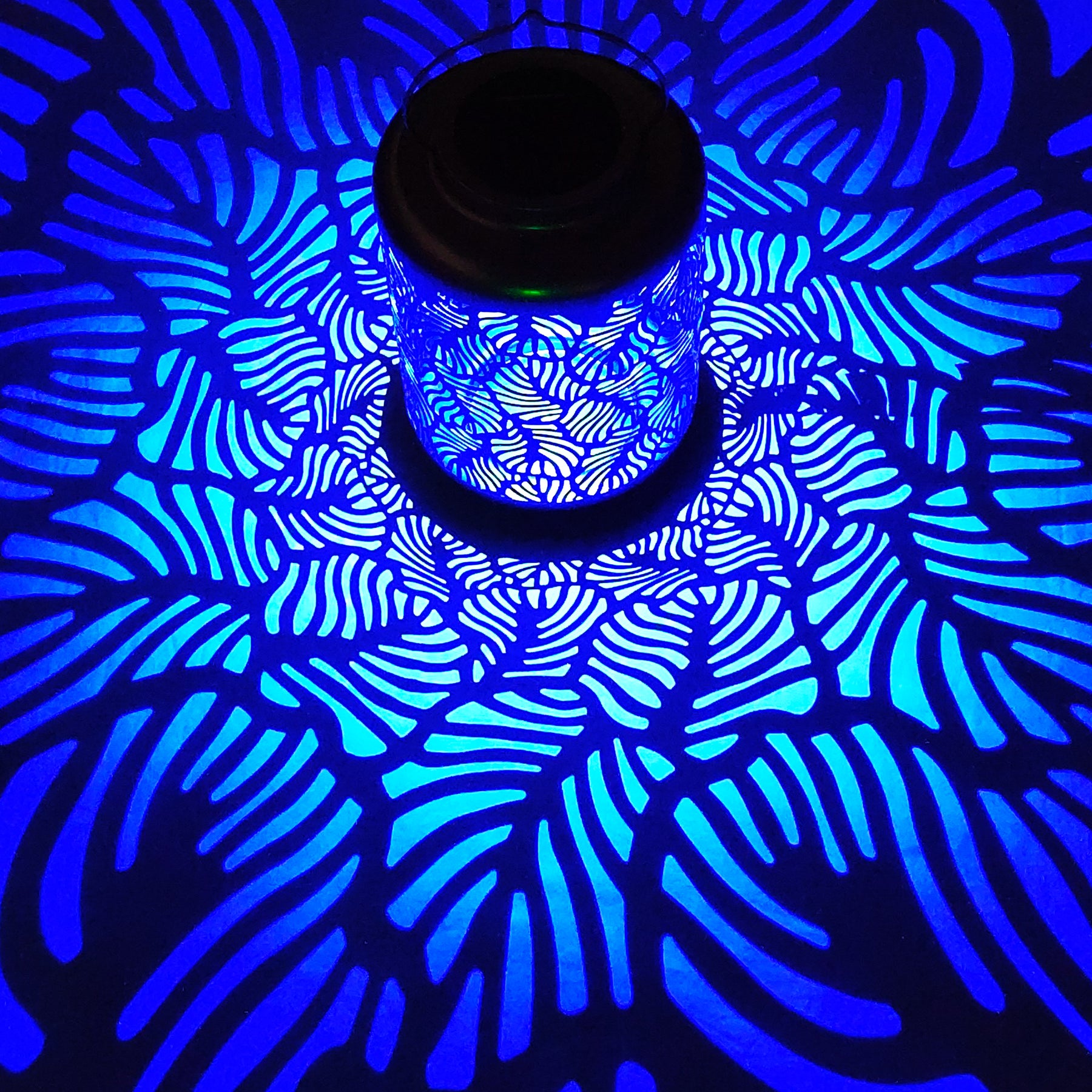 Bliss Outdoors 12-inch Tall Hanging and Tabletop Decorative Solar LED Lantern with Unique Tropical Leaf Design turned on and creating a cool pattern of blue light on the surface around it.