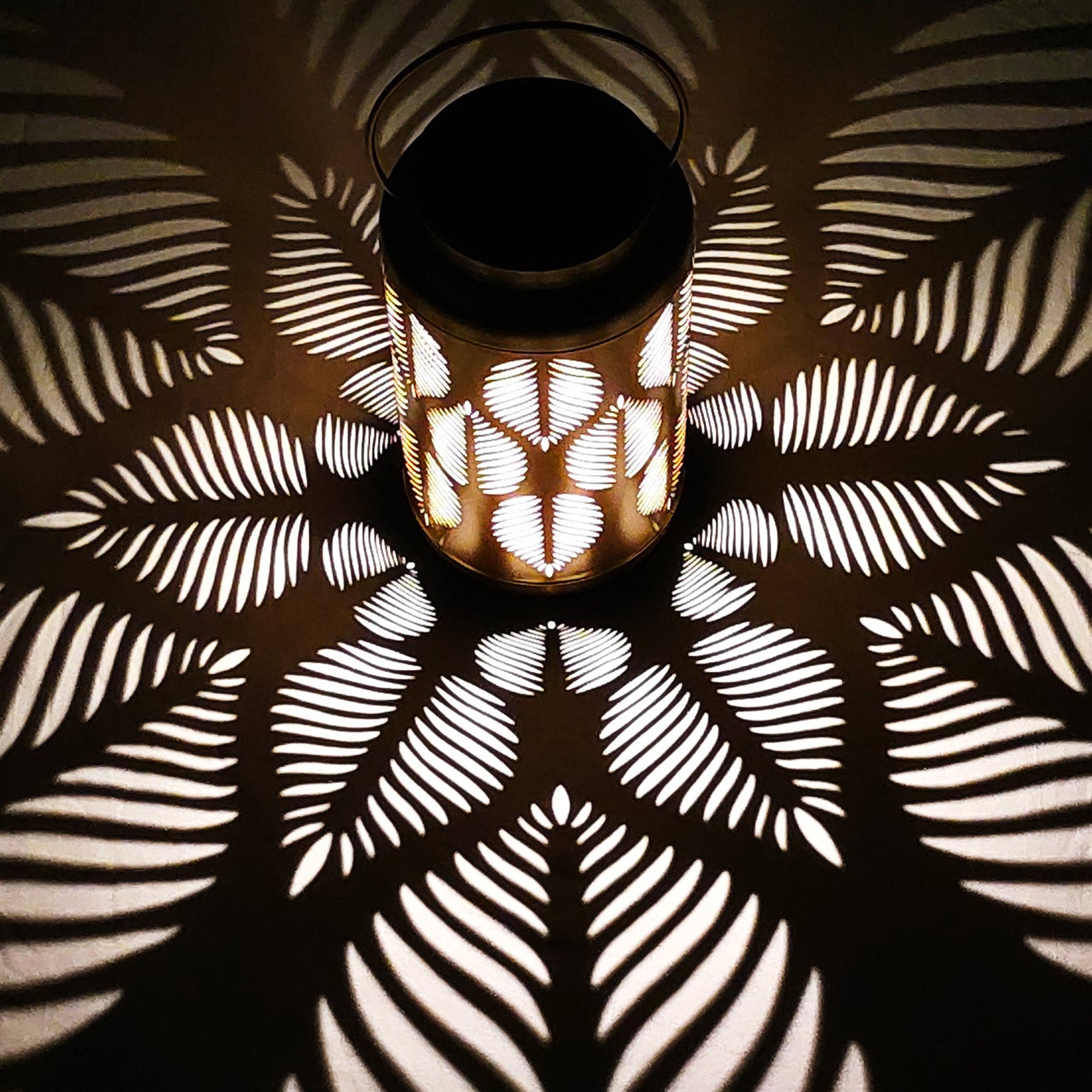 Bliss Outdoors 9-inch Tall Hanging and Tabletop Decorative Solar LED Lantern with Unique Tropical Leaf Design and Antique Hand Painted Finish turned on creating a cool light pattern on the surface around it.