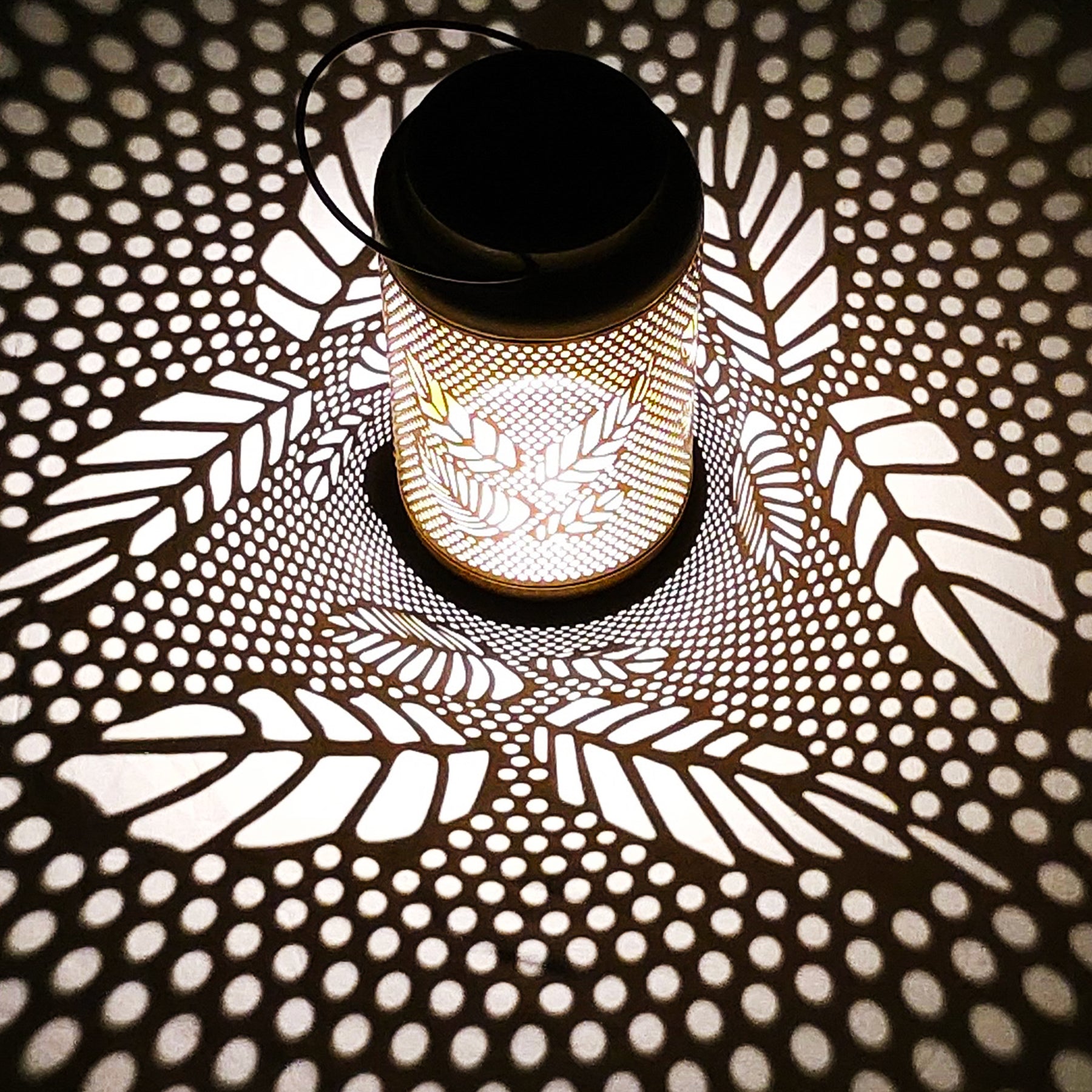 Bliss Outdoors 9-inch Tall Decorative Lantern with a berry leaf design turned on and creating a cool light pattern on the surface around it.