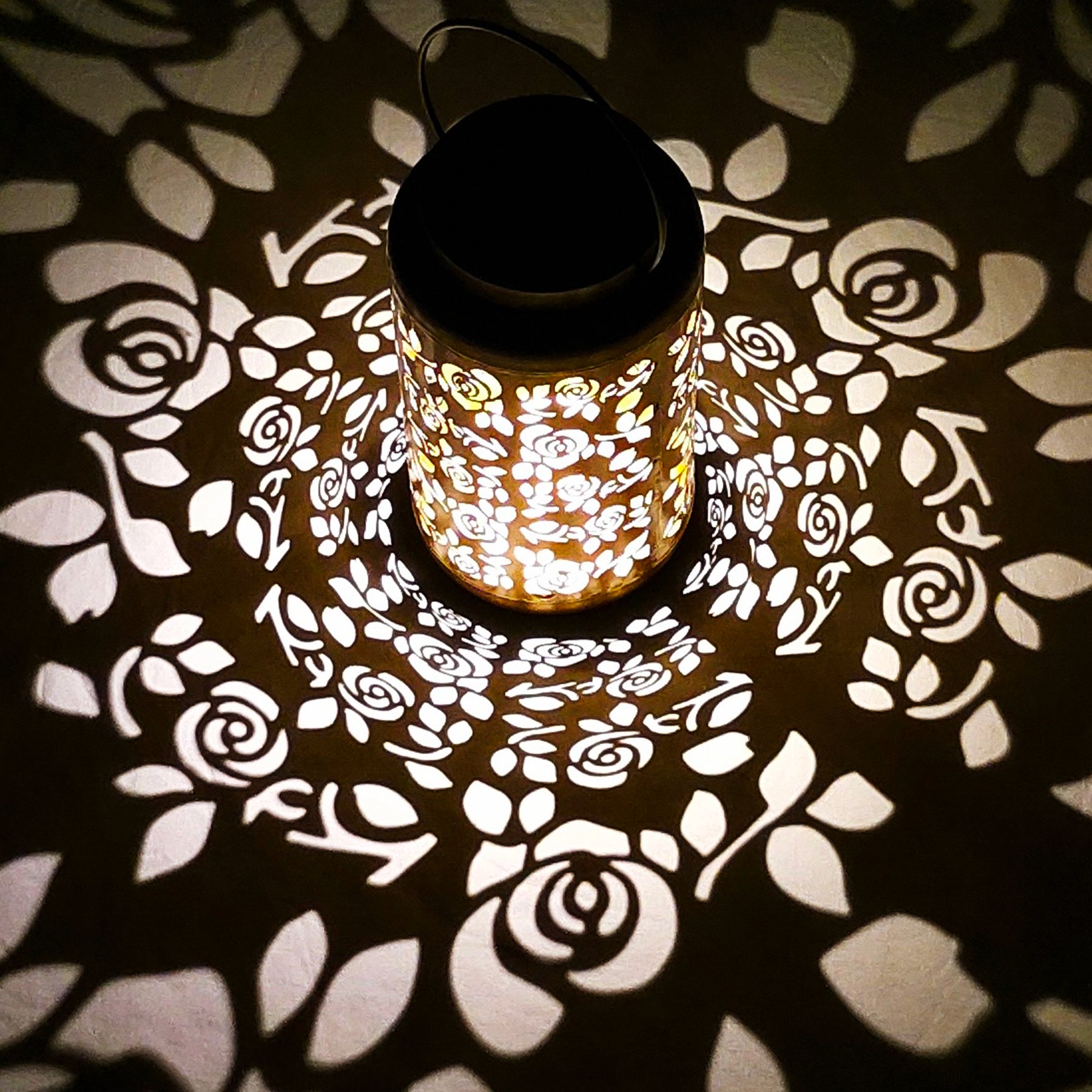 Bliss Outdoors 9-inch Tall Decorative Lantern with a unique rose design turned on and creating a cool light pattern on the surface around it.