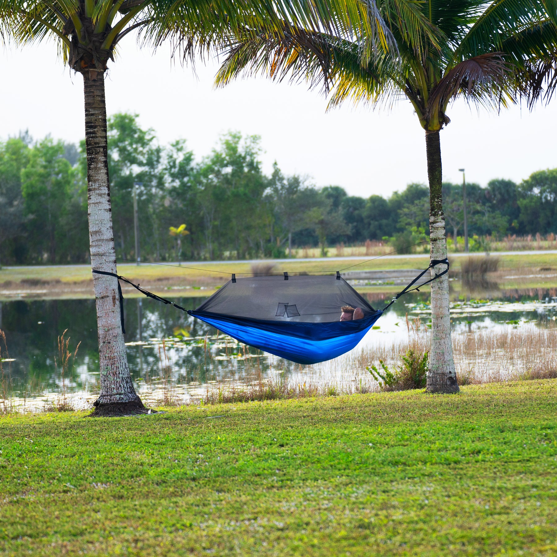 Person relaxing outside next to a body of water in a Bliss Hammocks 54-inch Wide Hammock in a Bag with mosquito net and tree straps between two palm trees.