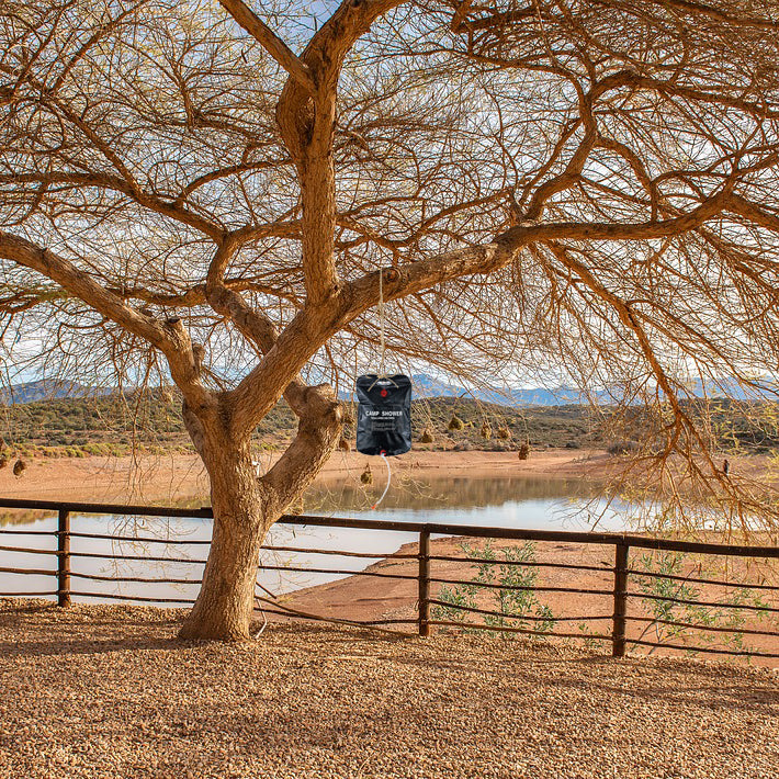 TrailGear 5 gallon Solar Shower Bag with Flexible Hose hanging from a tree branch outside.