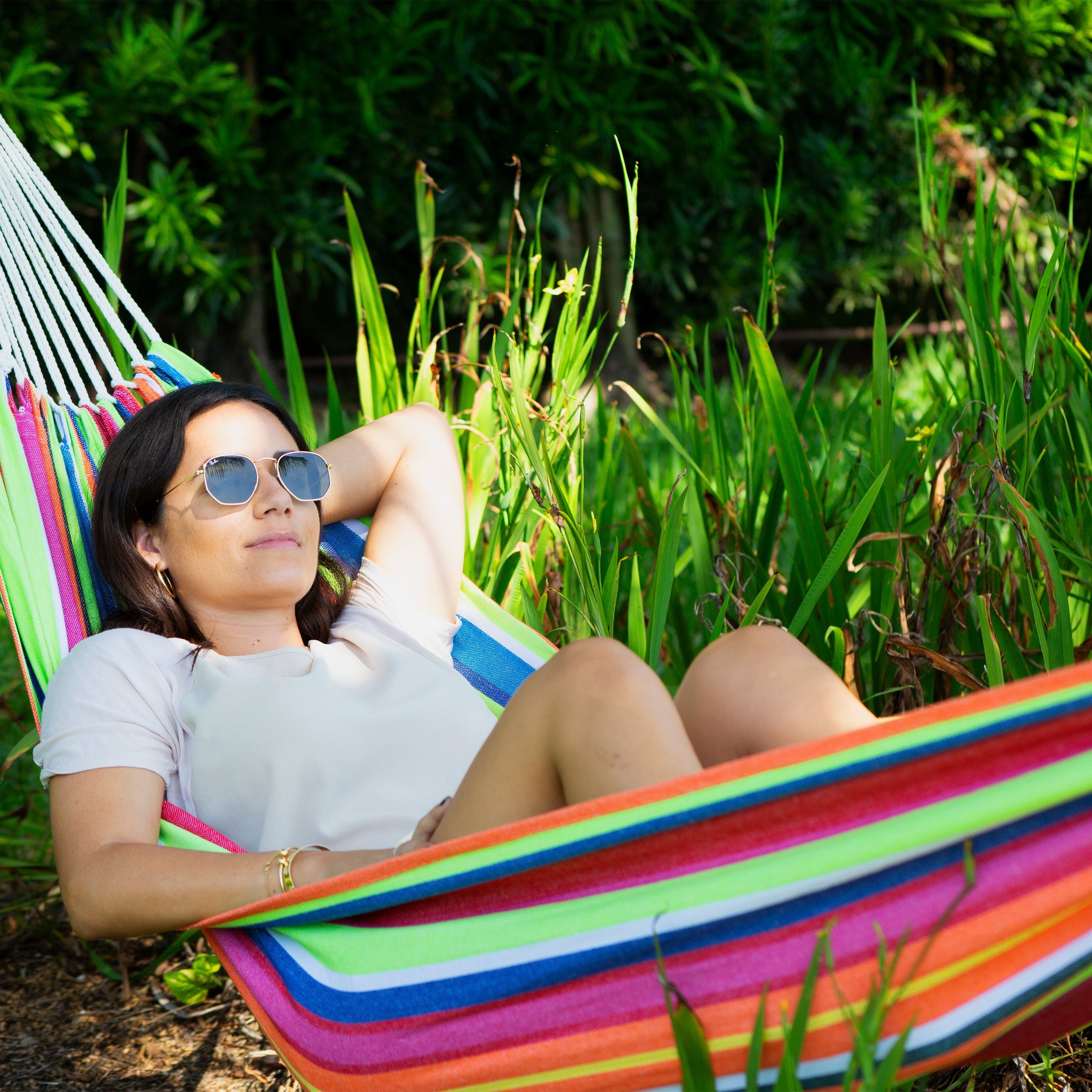 Woman relaxing in a colorful hammock from Bliss Hammocks. She's wearing sunglasses and surrounded by nature - Deals