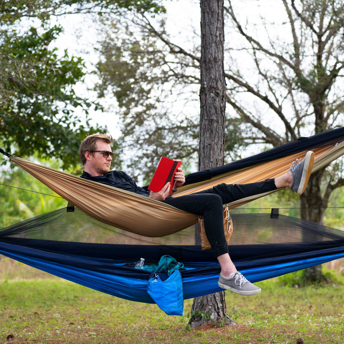 A man reading a book while relaxing in a Bliss Hammock Camping Hammock strapped between two trees. Another camping hammock is set up underneath him - Camping Hammocks