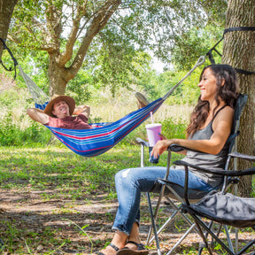 Man relaxing in a Bliss Hammocks 40-inch Wide Hammock hung outside between 2 trees. He is smiling at a woman in front of him sitting in a camping chair.