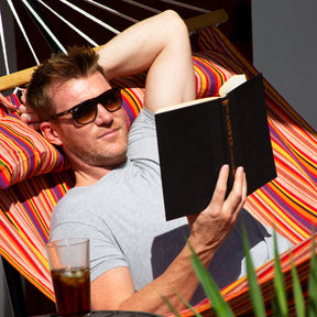 Man wearing sunglasses is reading a book while relaxing in a Bliss Hammocks 48-inch Wide Caribbean Hammock with a drink beside him on a side table.