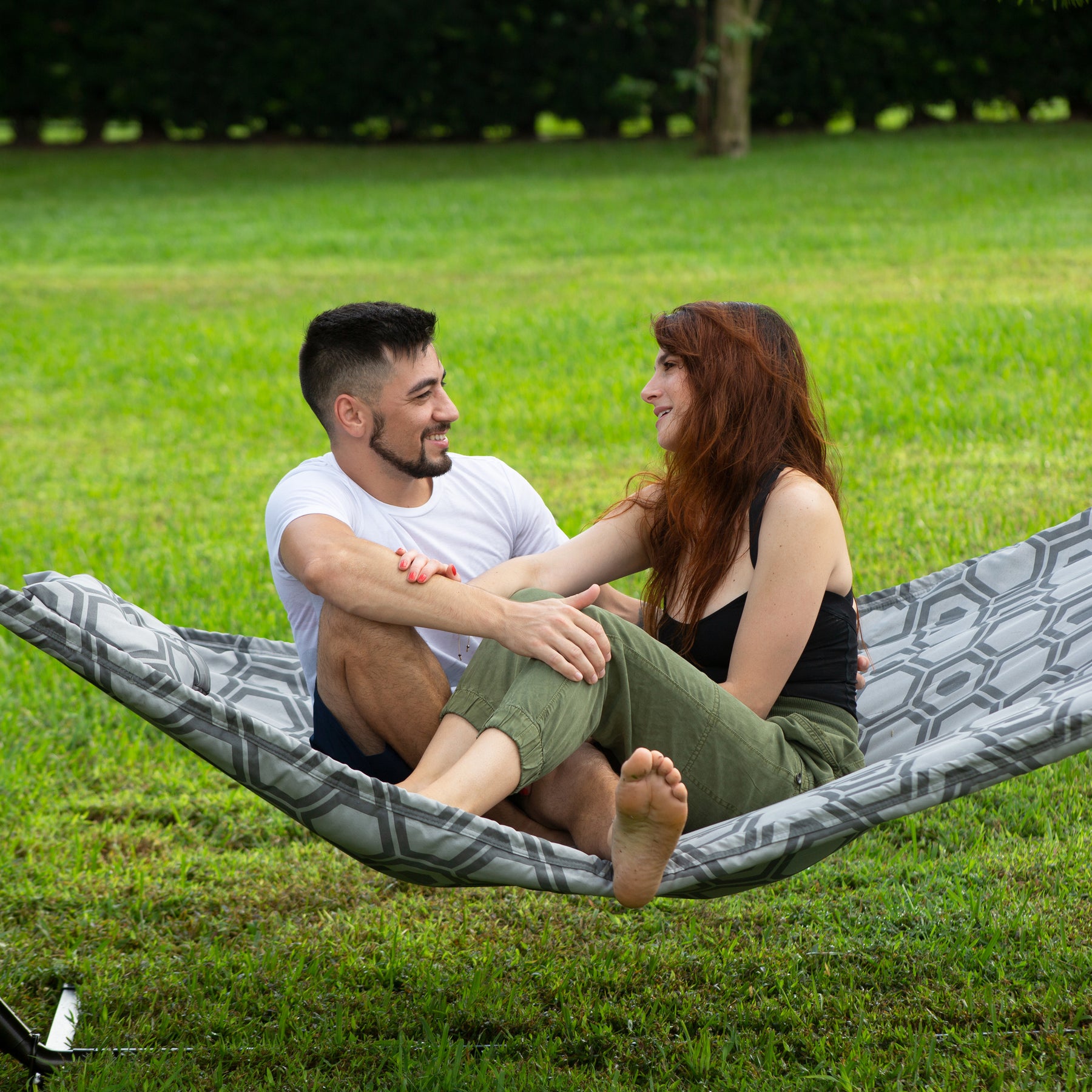 Man and woman sitting outside on the Bliss Hammocks 55-inch Wide 2-Person Reversible Quilted Hammock with spreader bars and a pillow.