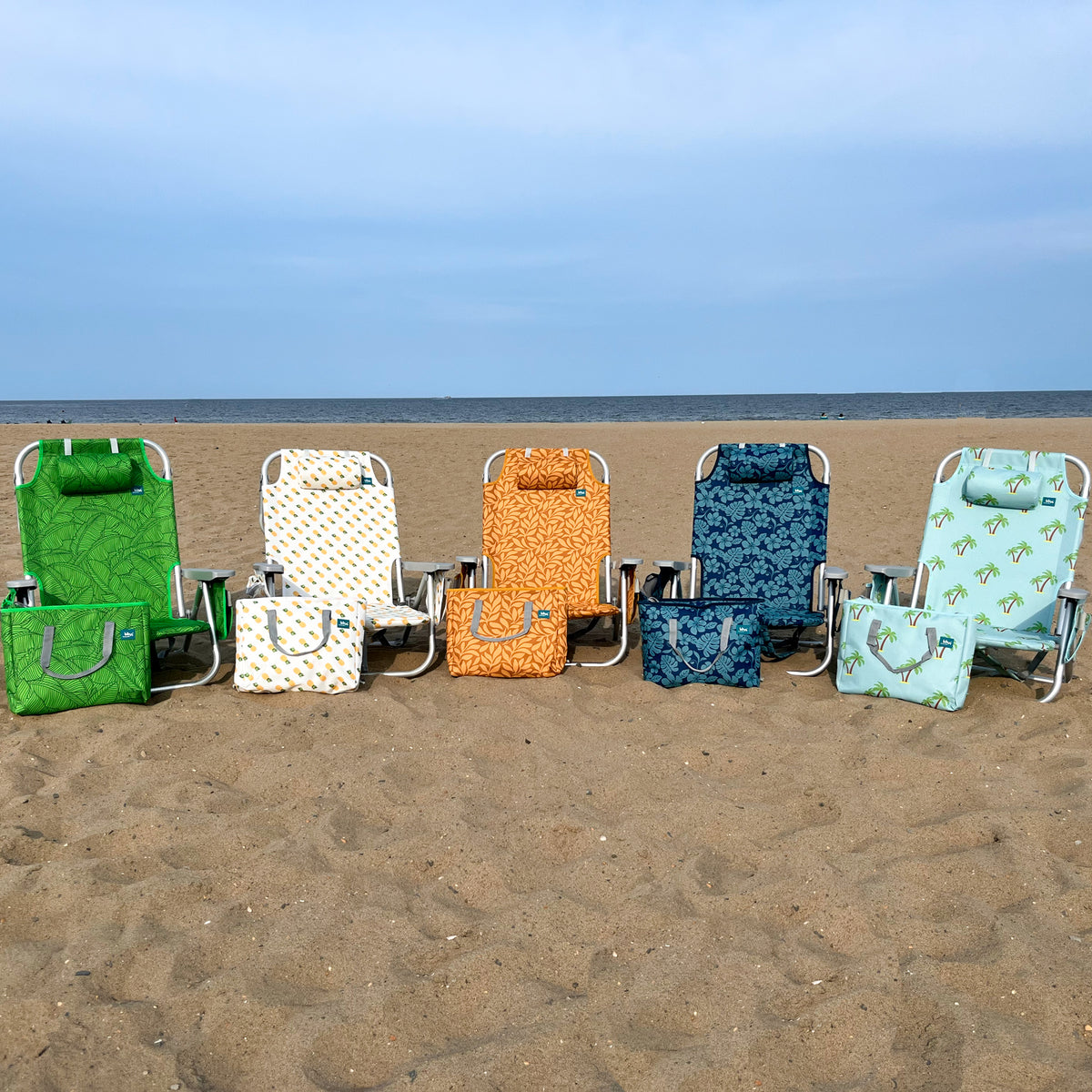 5 Backpack Aluminum Beach Chairs with different colors and patterns in a line on a beach with their matching cooler bags.
