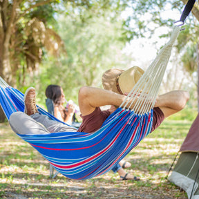 Man relaxing in a Bliss Hammocks Hammock between tree outside. He appears to be camping with a woman with a part of a tent shown and nature in the background.