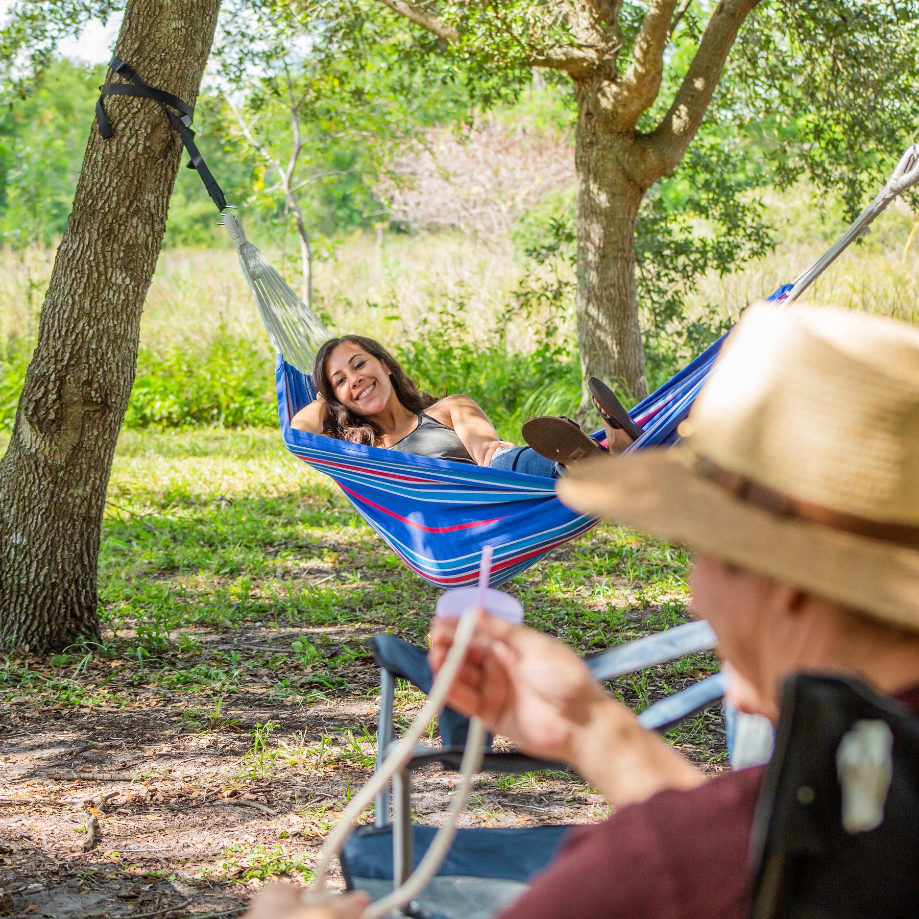 A woman relaxing in a Bliss Hammocks Hammock between trees outside. She is smiling towards the camera at a man in the foreground.