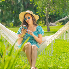 Woman using her phone while sitting in a Bliss Hammocks fringed hammock outside in the grass.