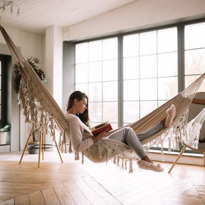 Woman reading a book in a Bliss Hammocks fringed hammock inside a house.