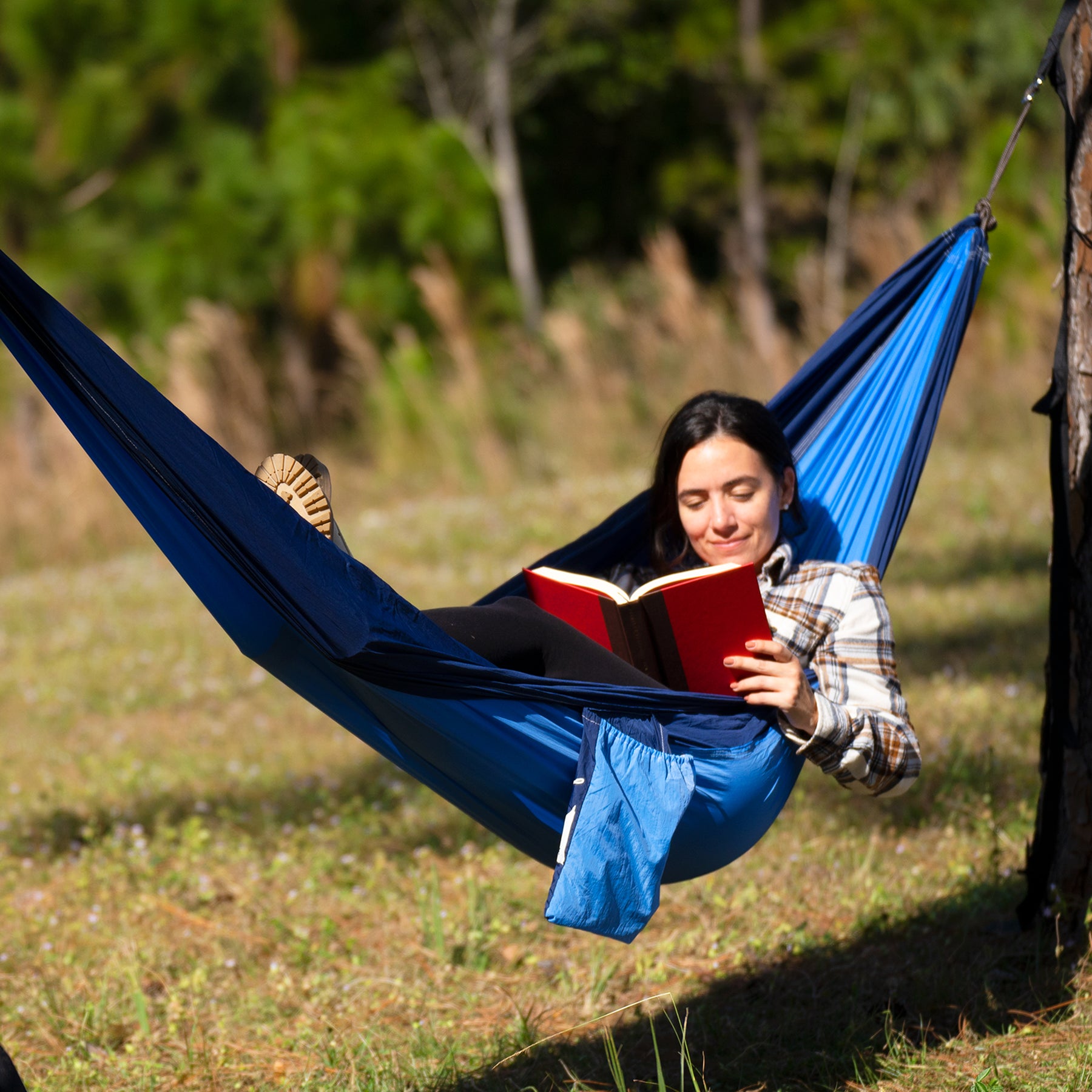 Woman reading a book while laying outside in the Bliss Hammocks 52-inch Wide Hammock in a Bag with Carabiners and Tree Straps in the royal bliss variation