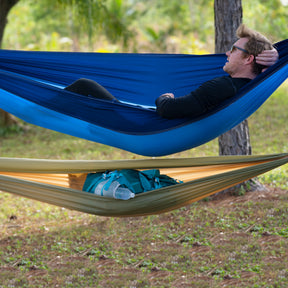 Man laying outside in the Bliss Hammocks 52-inch Wide Hammock in a Bag with Carabiners and Tree Straps in the royal bliss variation that is hung above another hammock.