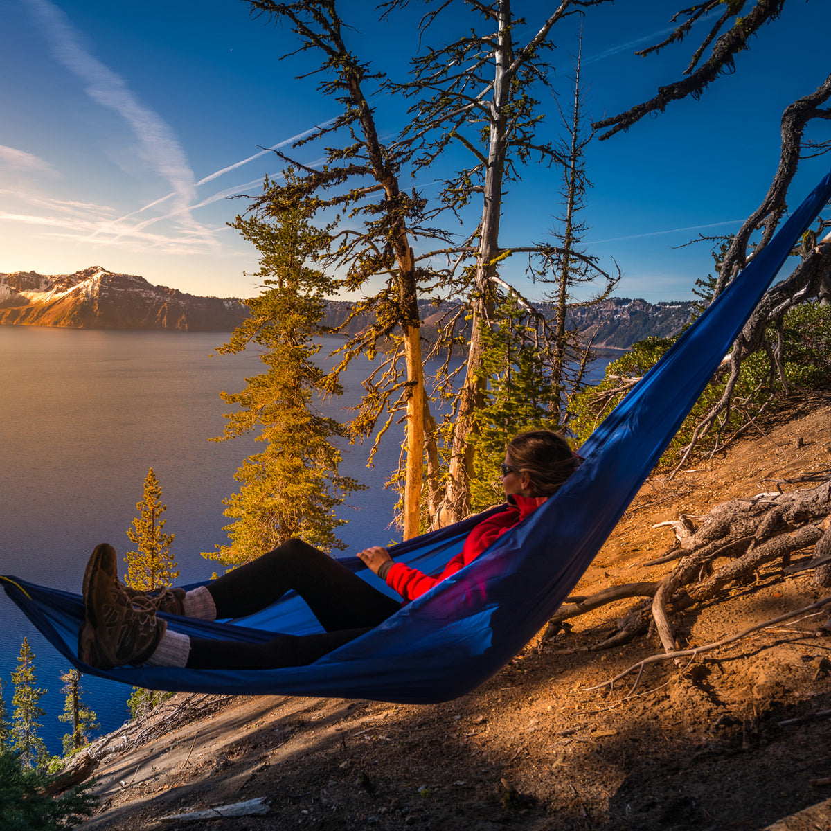 Woman relaxing outside in a Bliss Hammocks 54-inch Extra Wide To Go Hammock in a Bag next to a lake on the edge of a cliff. It's a clear day with mountains in the background.