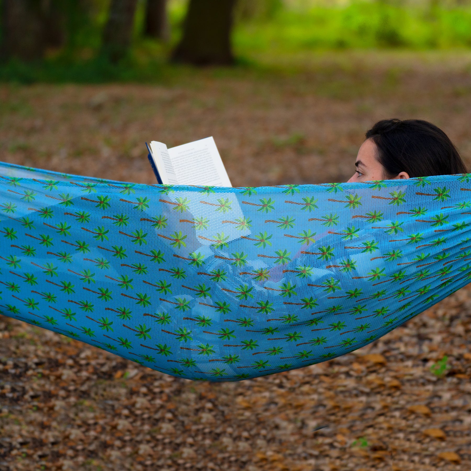 Person reading a book outside in the Bliss Hammocks 55-inch Mesh Hammock with a palm tree pattern.