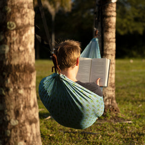 Man reading a book outside in the Bliss Hammocks 55-inch Mesh Hammock with a palm tree pattern hung between two trees.
