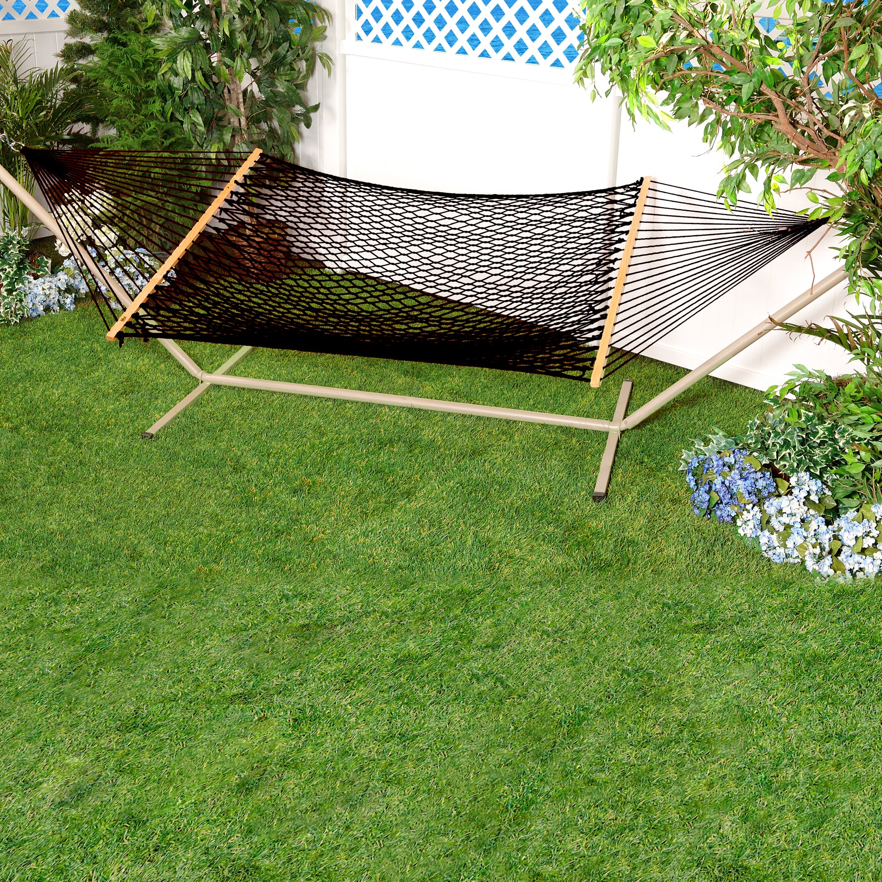Black Variation of Bliss Hammocks 60-inch Wide Cotton Rope Hammock with a stand in a yard by some flowers and a fence.