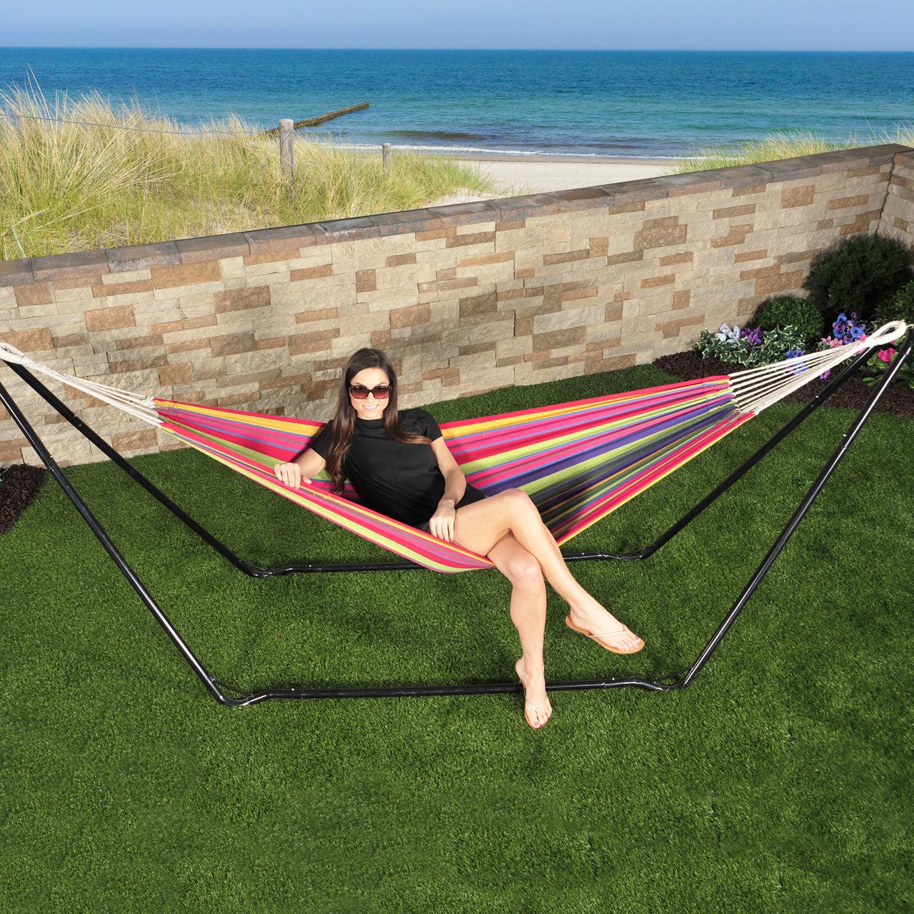 Woman sitting in a Bliss Hammocks Multi-Color hammock attached to the Bliss Hammocks 10-foot Hammock Stand with Hanging hooks. She is in an enclosed yard with the beach and the ocean in the background.