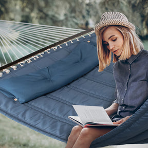 Girl reading a book while sitting in the Bliss Hammocks 55-inch Wide Quilted Hammock with Spreader Bars and Pillow in the denim blue variation.