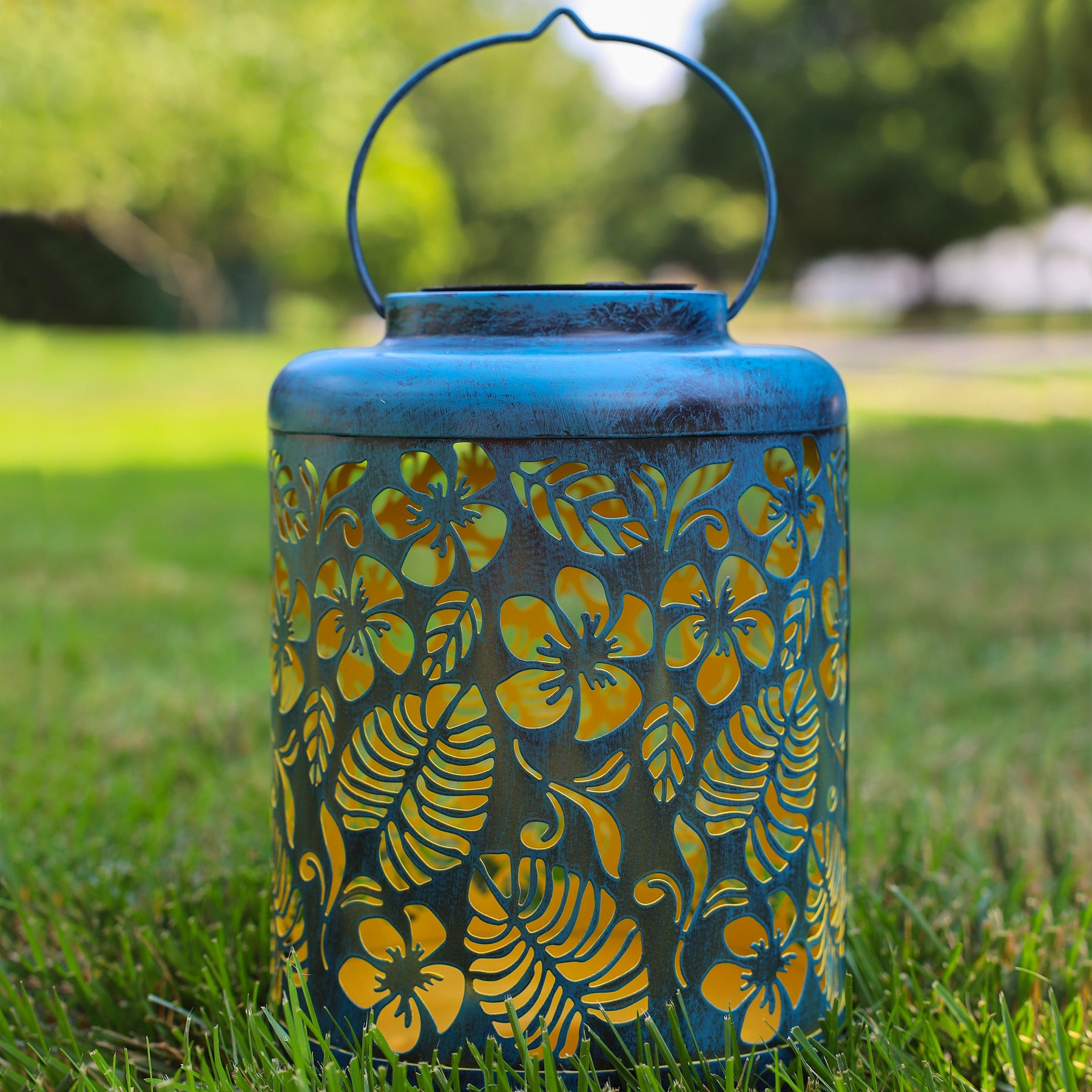 Blue Solar Lantern with tropical flower design standing on grass.