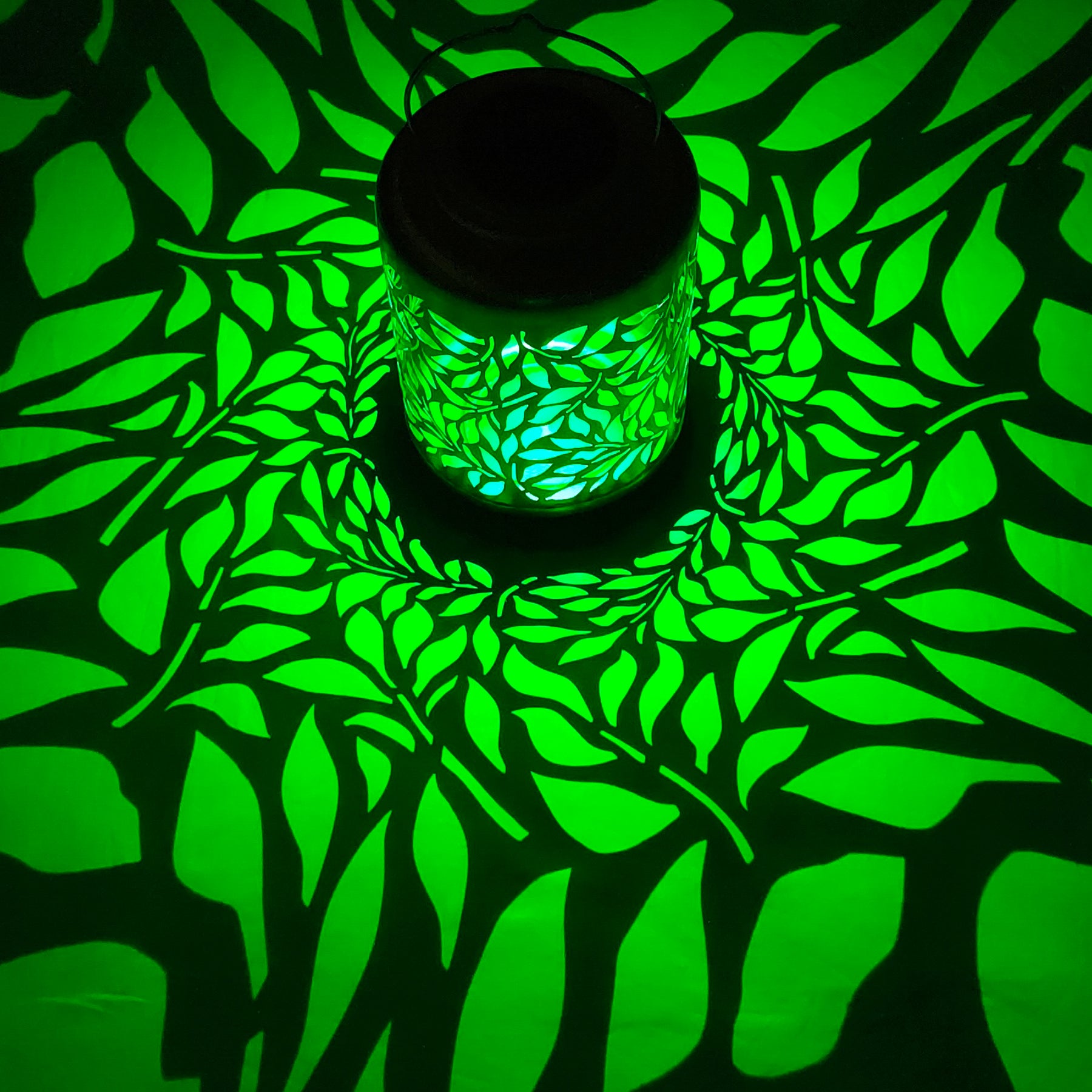 Bliss Outdoors 12-inch Tall Hanging and Tabletop Decorative Solar LED Lantern with Unique Olive Leaf Design turned on and creating a cool pattern of green  light on the surface around it.