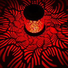 Bliss Outdoors 12-inch Tall Hanging and Tabletop Decorative Solar LED Lantern with Unique Tropical Leaf Design turned on and creating a cool pattern of red light on the surface around it.