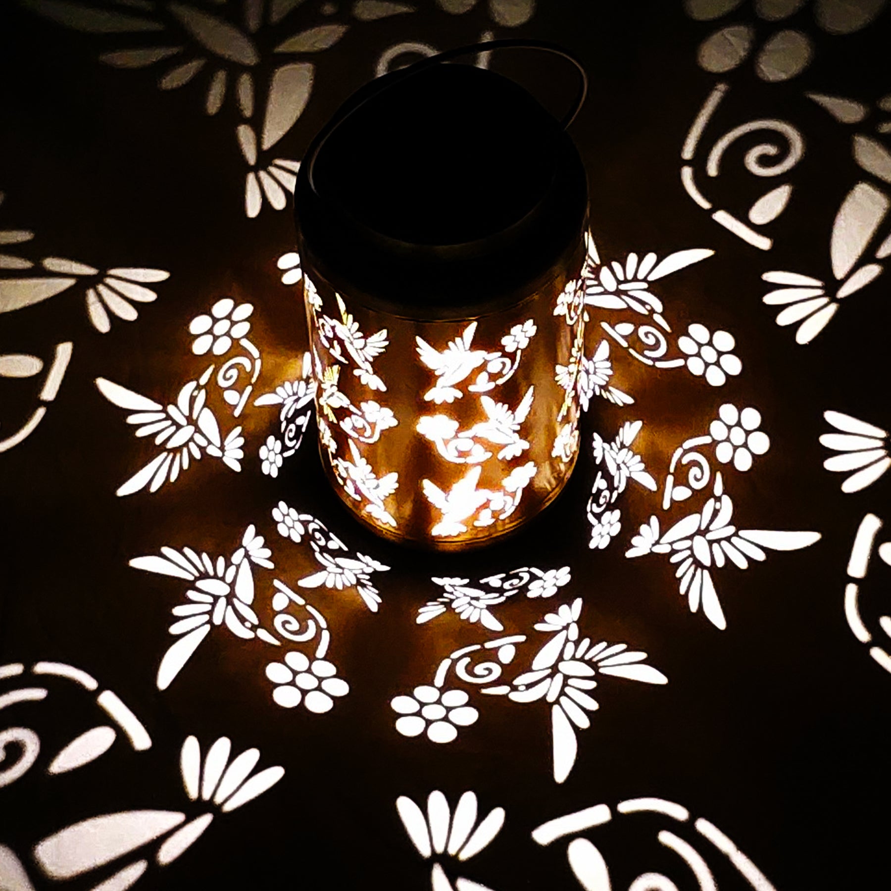 Bliss Outdoors 9-inch Decorative Solar LED Lantern with Unique Humming Bird Design turned on and creating a cool light pattern on the surface around it.