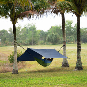 Person relaxing in a hammock hung between palm trees with the Bliss Hammocks Rain Shelter secured above him.