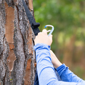 Person using a carabiner to hook on the tree strap that is tied around a tree.