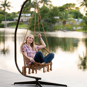 Woman sitting in the Bliss Hammocks 31.5-inch Wide Brown Macramé Swing Chair next to a river.