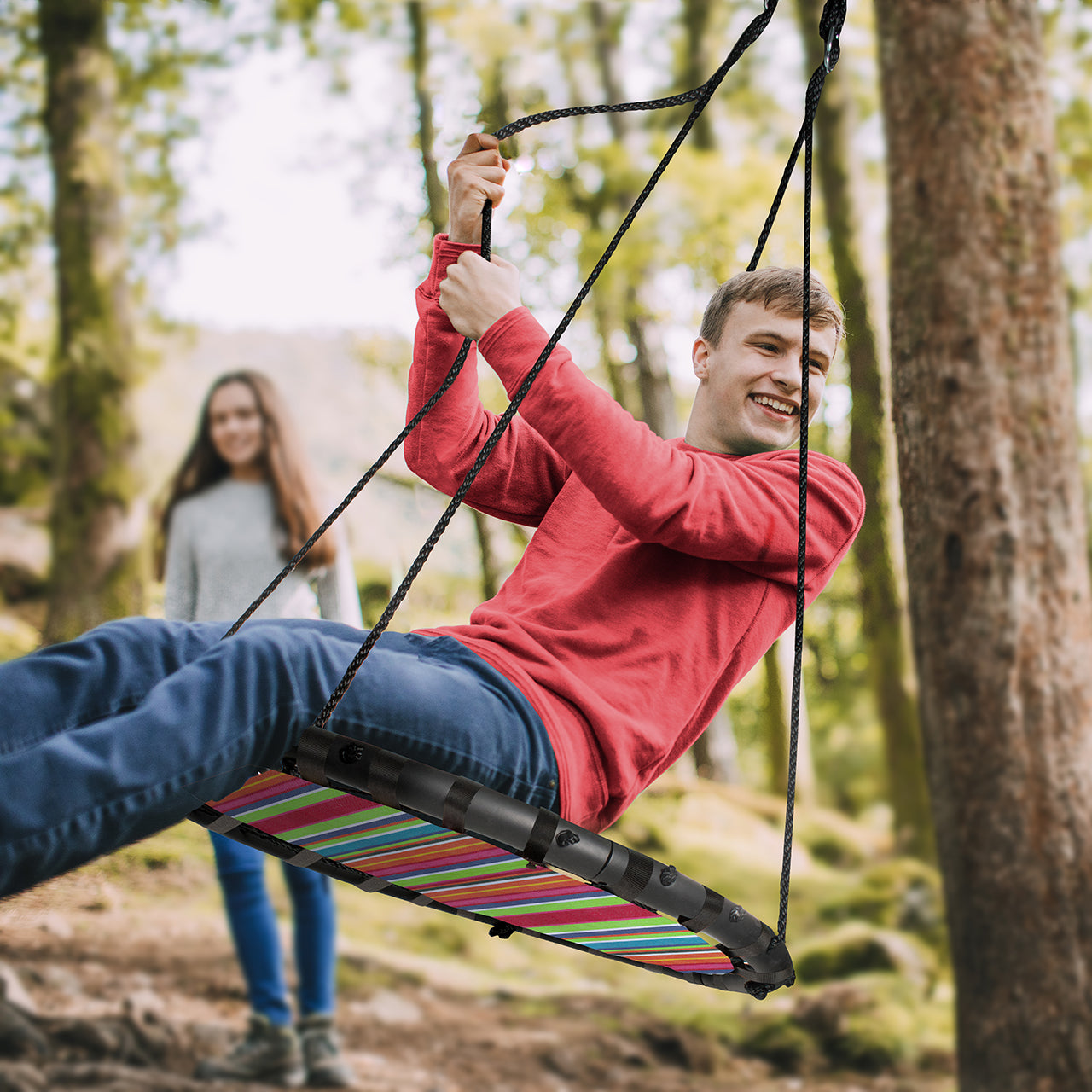 Boy swinging from a tree on the Bliss Outdoors Striped Fabric Tree Glider. A girl in the background is watching him have fun.