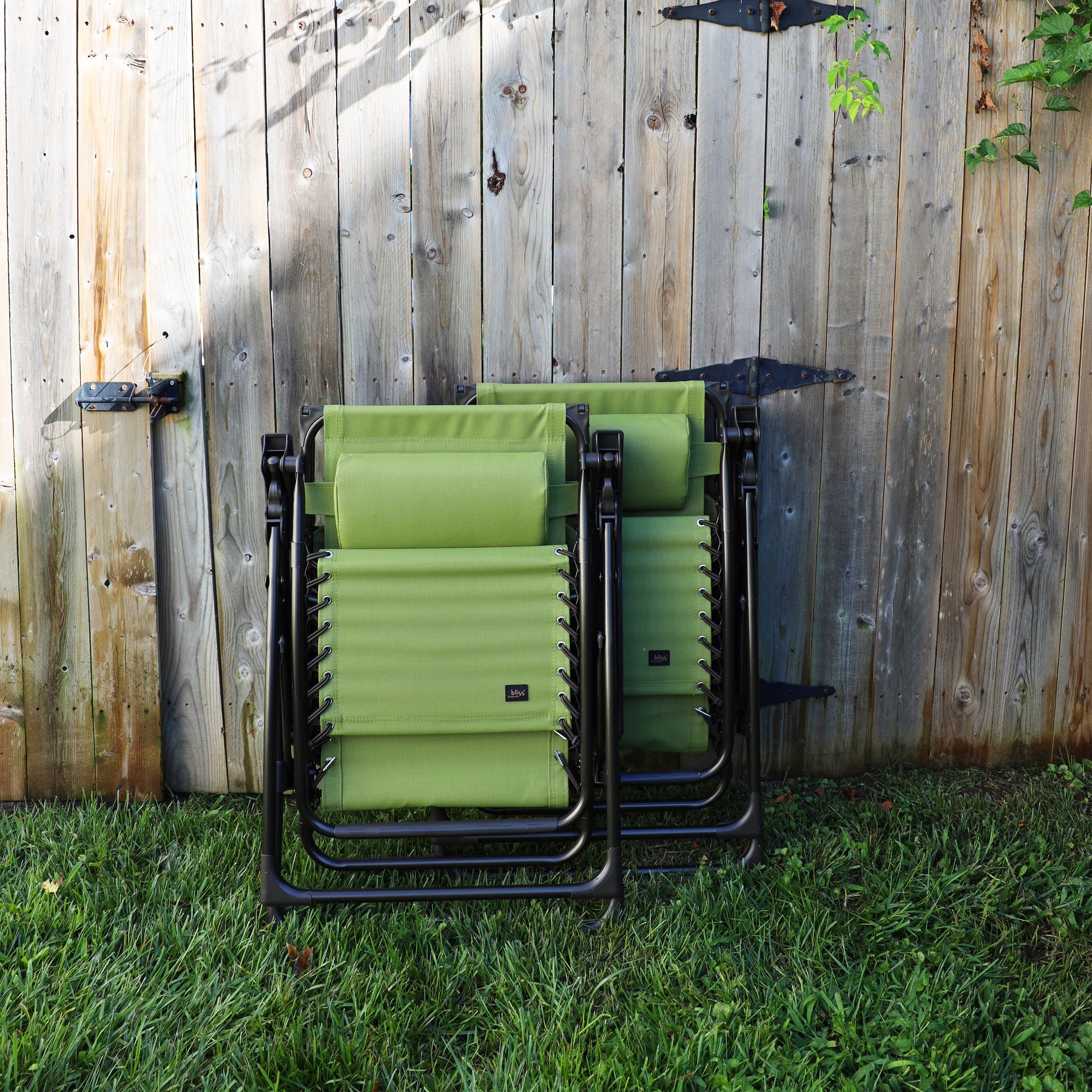 Set of 2 26-inch sage green Zero Gravity Chairs folded and leaning against a fence.