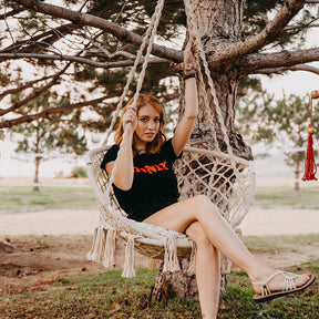 Girl sitting in the Bliss Hammocks 31.5-inch Wide Macramé Swing Chair with Fringe lining and Padded Cushion that is hanging from a tree outside.
