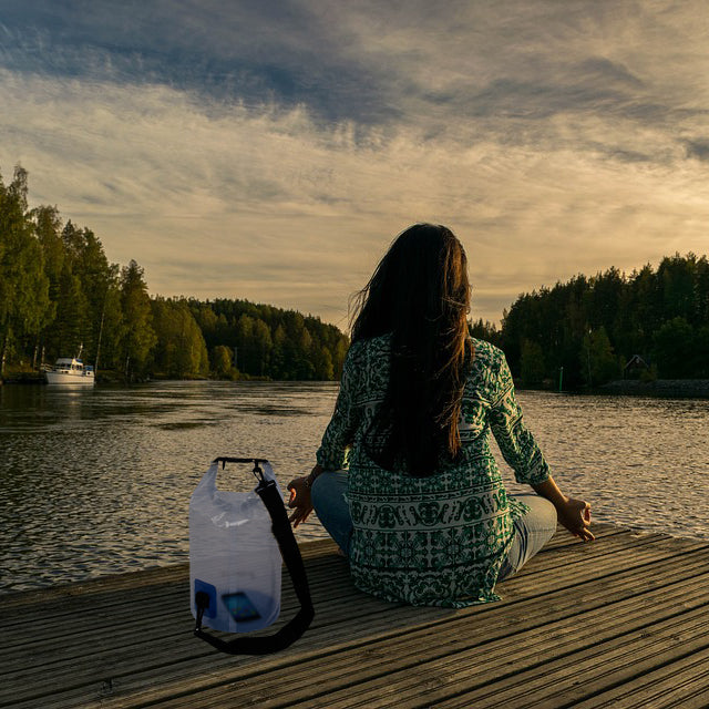 Woman meditating on a dock overlooking a lake with a TrailGear 5 liter Heavy-Duty Transparent Dry Bag beside her.