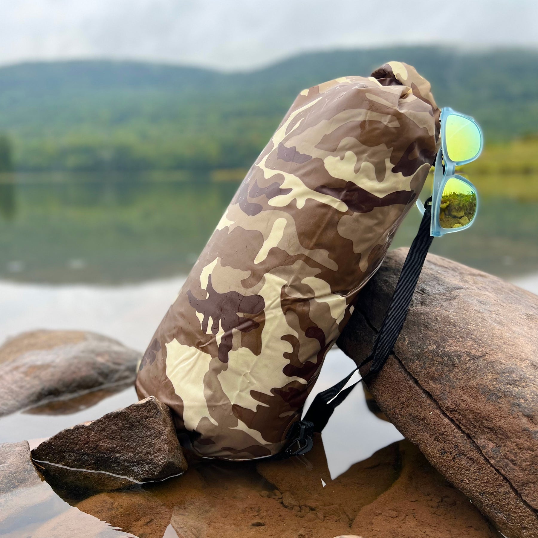 TrailGear 10 liter Heavy-Duty Camouflaged Dry Bag in the brown camo variation in shallow water and leaning against a rock with sunglass hung on the side of the bag.