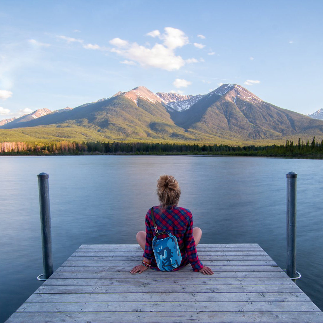 Girl sitting on a dock overlooking a lake and mountains in the background. She has a TrailGear 5 liter Heavy-Duty Camouflaged Dry Bag in the blue camo variation strapped to her back.